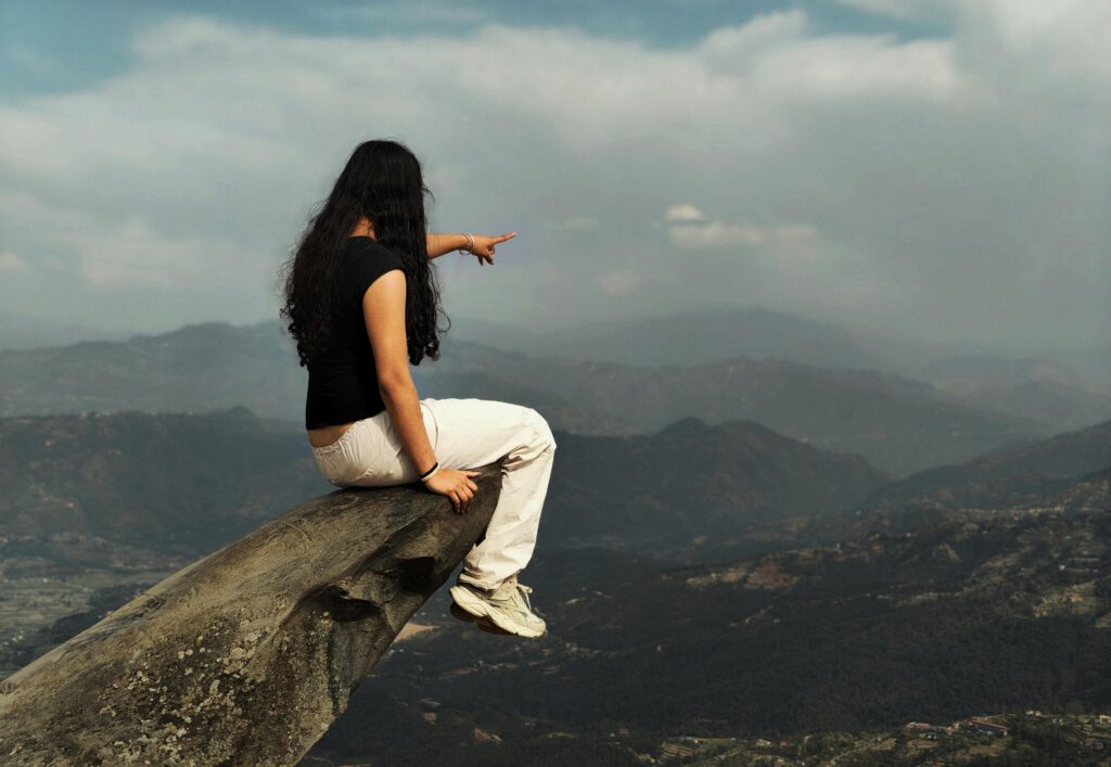 Woman sitting on a rock ledge, pointing at the scenic mountainous landscape.