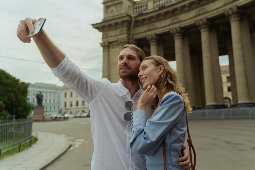 Couple taking a selfie in front of historic columns on a sunny day, capturing a happy moment.