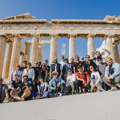 A group photo of tourists in front of the Parthenon, Athens, Greece under a clear blue sky.